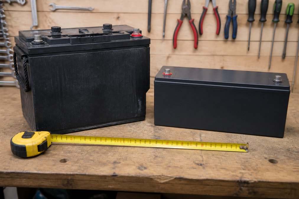 Close-up of a large, heavy AGM lead acid leisure battery sitting on a workshop bench next to a slim lithium LiFePO4 battery of the same capacity, with a tape measure and workshop lighting highlighting the size and weight difference