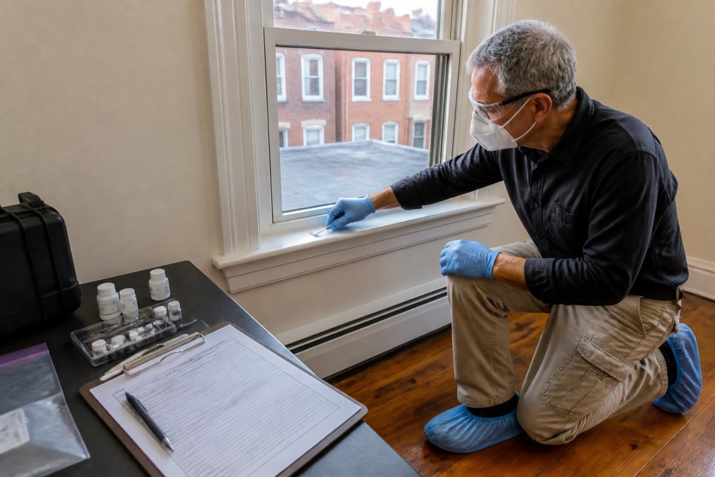 Certified lead inspector conducting dust wipe sampling test on a windowsill in an older residential property, with testing equipment, sample containers, and compliance documentation visible on a nearby table