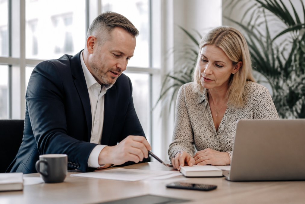 A business coach in a one-on-one session with a small business owner at a conference table, both reviewing notes and a laptop, bright modern office setting