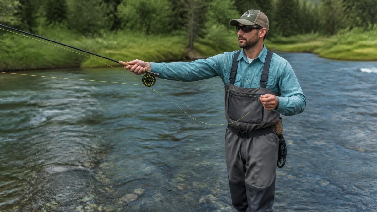 Professional fly fishing guide demonstrating proper casting stance and grip with a fly rod while standing in shallow river water, showing arm position and body alignment with clear water backdrop