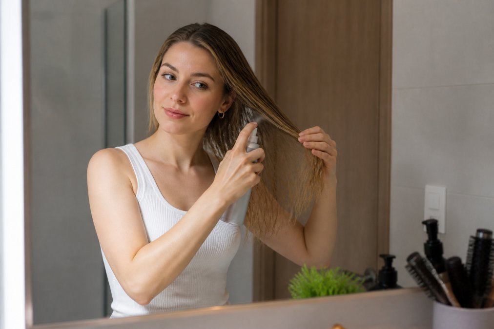 Person with fine hair applying heat protectant spray before blow-drying, showing proper application technique from mid-length to ends
