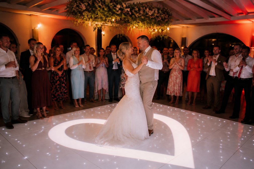 Bruidspaar danst op een verlichte dansvloer met een hartpatroon, omringd door juichende gasten, warme up-lights baden de feestruimte in zachte tinten
