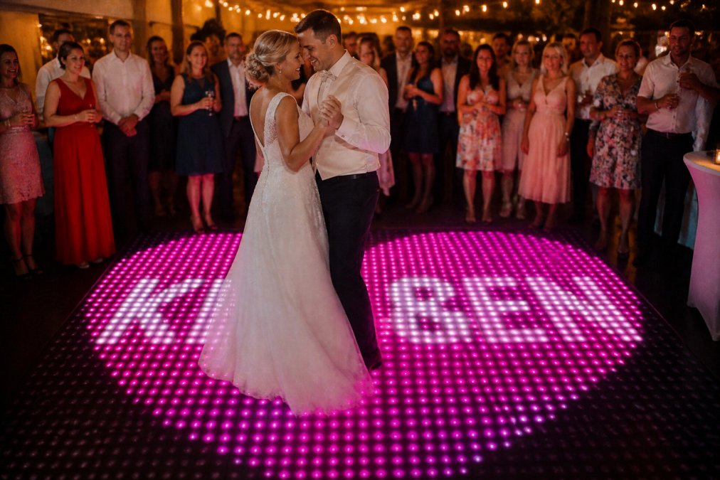 Bruidspaar dansend op een verlichte LED dansvloer met twinkle effect, omringd door gasten die toekijken, warm gouden licht, feestelijke sfeer