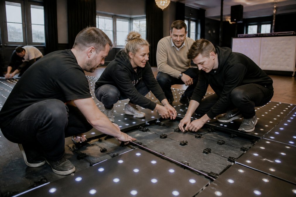 Groep mensen dansend op een volledig verlichte LED-dansvloer in een feestzaal, RGB-kleuren zichtbaar, vrolijke bruiloftssfeer met bruid en bruidegom, Petra Keizer zichtbaar in beeld