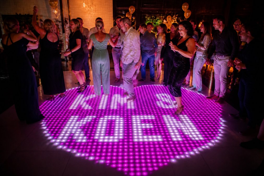 Groep mensen dansend op een helder verlichte dansvloer tijdens een feest, met kleurige LED-effecten zichtbaar onder hun voeten, gekleed in feestkleding en duidelijk plezier hebbend