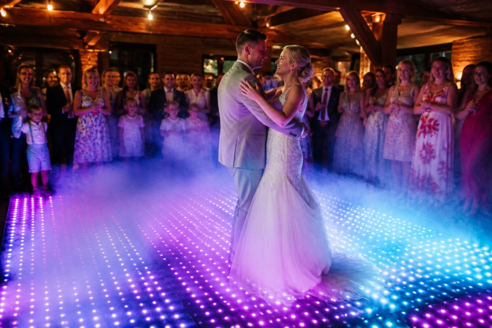 Bruidspaar dansend op een verlichte LED dansvloer met RGB-kleuren die gloeien in paars en blauw, gasten kijkend vanaf de zijkant, festive disco-effect met laaghangende rook, warmgelicht interieur