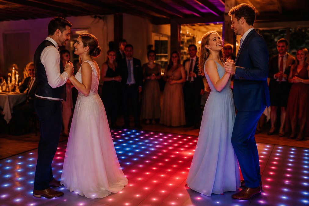 Twee stellen dansend op een verlichte dansvloer op een bruiloft, omringd door toekijkende gasten, warme rode en blauwe lichteffecten, feestelijke sfeer met kaarsen op tafels op de achtergrond
