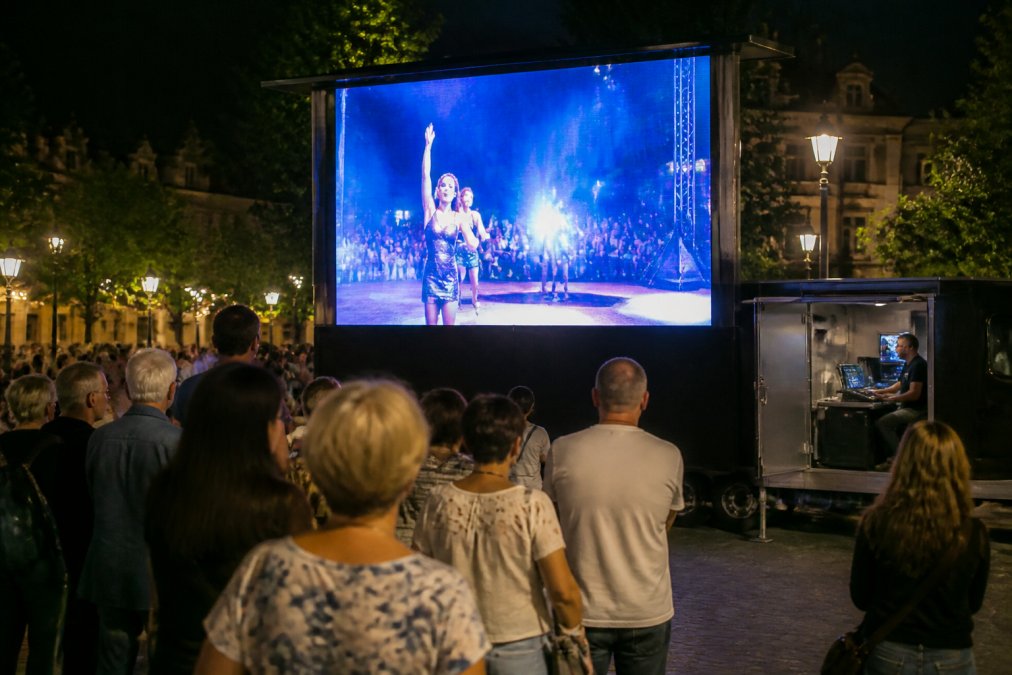Een mobiele LED-aanhangwagen met verlicht scherm op een stadsplein tijdens een outdoor evenement, mensen kijken ernaar in de avond, regiekamer zichtbaar achter het scherm