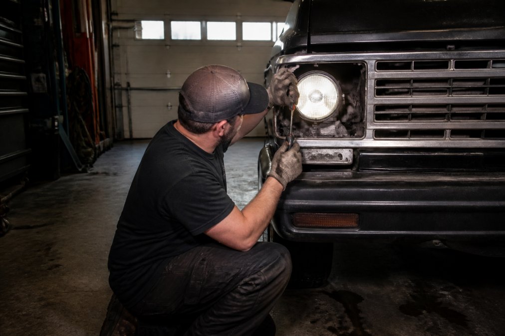 Mechanic using a screwdriver to adjust the headlight alignment of a classic truck against a garage wall