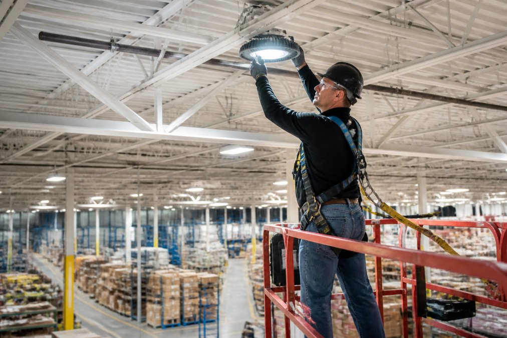 Professional technician in safety harness on elevated lift platform installing LED fixture in high-ceiling warehouse with industrial infrastructure visible below
