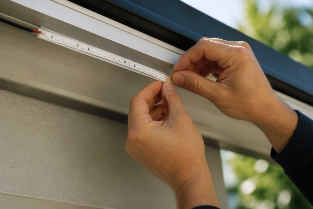 Professional's hands carefully peeling adhesive backing from an LED strip light and pressing it firmly onto a clean aluminum mounting channel in natural daylight