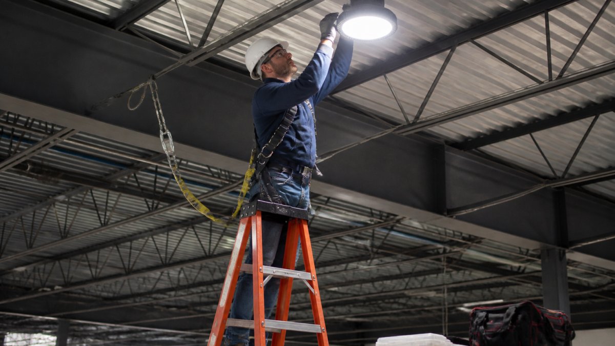 Professional technician installing LED fixtures in high-bay warehouse ceiling wearing safety harness, with ladder and professional installation equipment visible, showing scale and complexity of industrial lighting installation