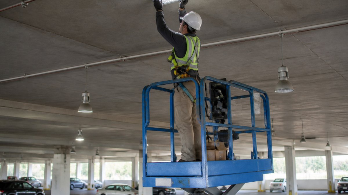 Professional technician on a hydraulic lift installing LED retrofit fixtures in a high-bay parking garage, with older HID fixtures visible in the background and structural concrete pillars below