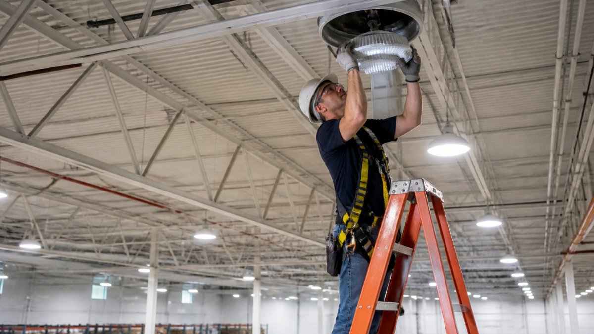 Professional technician in safety harness installing LED retrofit kit into recessed housing in high-bay warehouse ceiling, with ladder and safety equipment visible