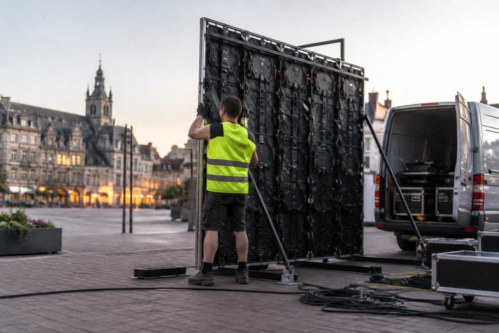 Een technicus in veiligheidshesje stelt een LED-paneel van ongeveer 3 bij 3 meter op aan de rand van een openbaar plein in een Vlaamse stad, met een bestelwagen en kabels op de achtergrond bij zacht avondlicht