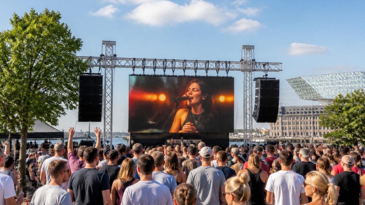 Live evenement in volle gang met groot LED-scherm in werking, publiek kijkt naar de beelden op een zonnige dag, professionele truss-constructie zichtbaar, energieke sfeer met mensen die genieten van het scherm