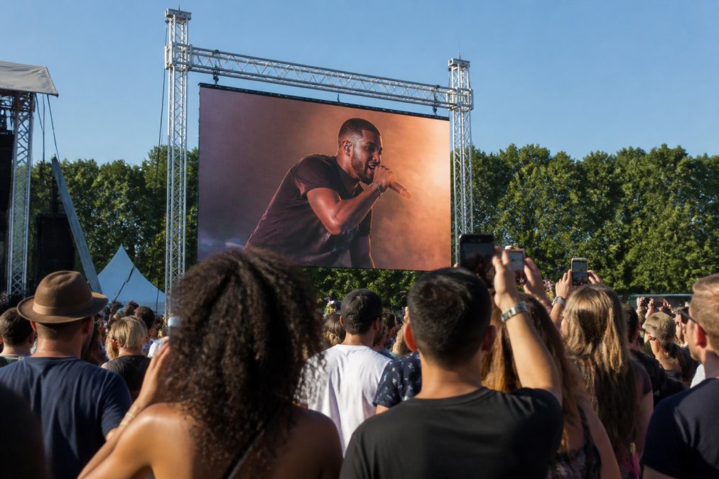 Groot LED scherm op outdoor festival met publiek dat ernaar kijkt in fel daglicht, bomen op achtergrond, bezoekers fotograferen de schermen