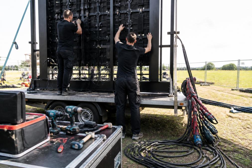 Technische medewerkers die modulair LED scherm monteren op aanhangwagen, elektrische kabels zichtbaar, gereedschap op tafel, daglicht in achtergrond