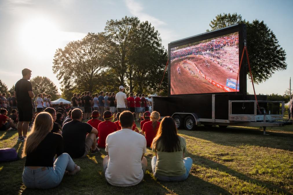Mobiel LED-scherm op aanhangwagen gepositioneerd op een openlucht evenement met bezoekers die naar het scherm kijken en de zon die licht werpt op de aanhangwagen