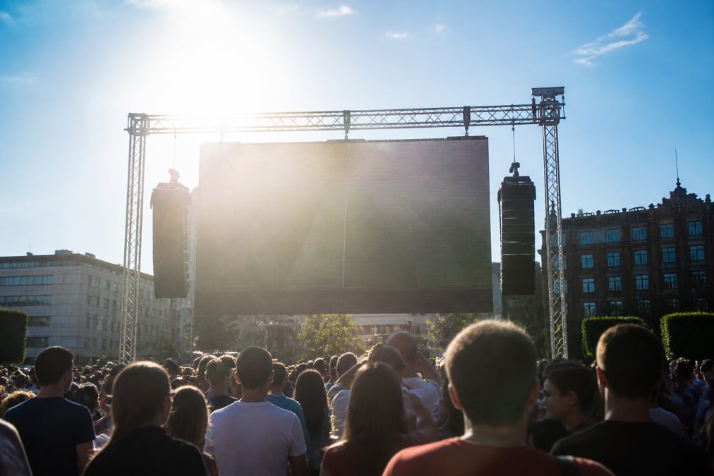 Outdoor evenement in volle zon met groot LED-scherm zichtbaar tegen heldere hemel, bezoekers kijken naar het display, fel zonlicht valt direct op het schermoppervlak