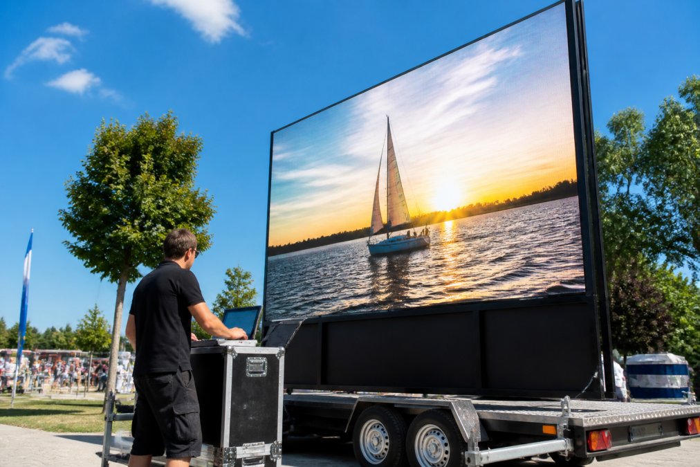 Professionele technicus controleert instellingen van groot LED-scherm op aanhangwagen tijdens outdoor evenement, scherm toont helder beeld in daglicht, zonnig weer