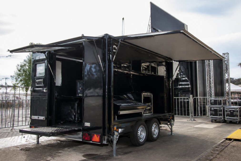 Mobiele LED aanhangwagen geparkeerd op evenementenlocatie met zijkanten open, regieruimte zichtbaar met monitoren en apparatuur, professioneel opgesteld tegen achtergrond van evenemententerrein