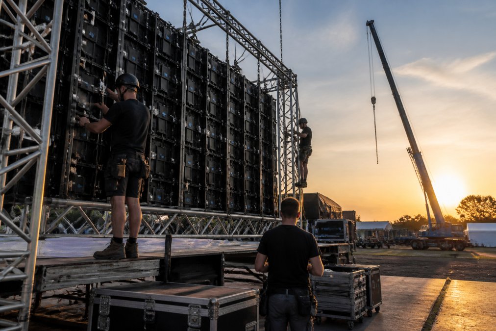 Technisch team monteert een modulair LED-scherm op een truss-constructie op een evenementenlocatie, kabels netjes weggewerkt, late namiddagzon, hijskraan op de achtergrond