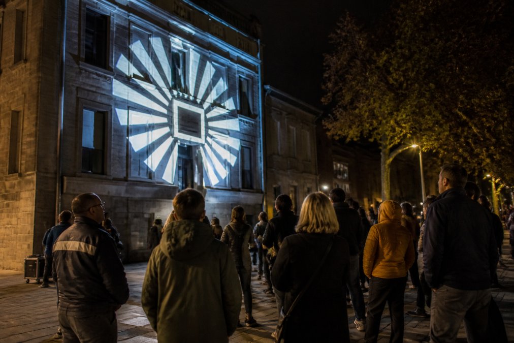 Avondscène met lichtprojectie van een logo op de gevel van een gebouw, met voorbijgangers die naar de projectie kijken, dramatische verlichting
