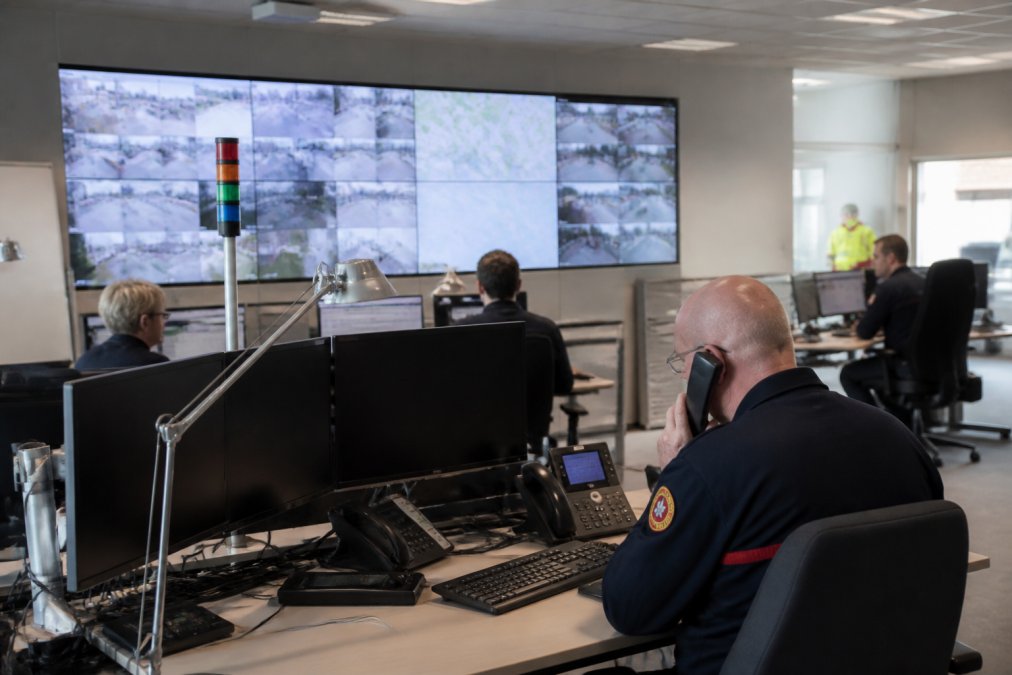 Gemeentelijk crisiscentrum in Antwerpen met medewerkers die realtime waarschuwingsberichten monitoren op grote schermen, controlekamer met meerdere displays en communicatieapparatuur, heldere verlichting en professionele werkstations