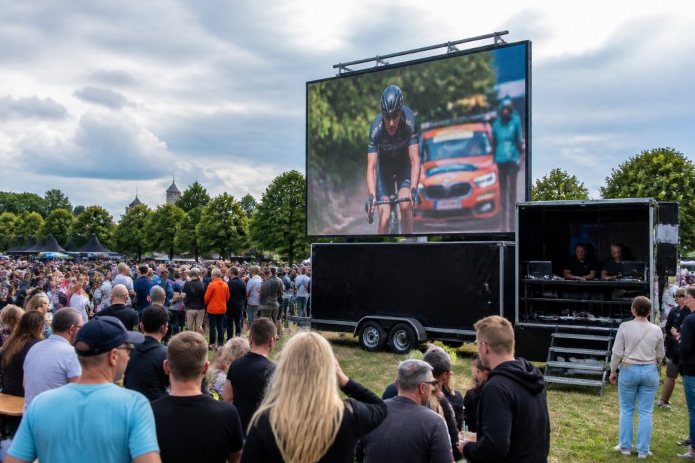 Mobiel LED-scherm op aanhanger geplaatst op een druk evenemententerrein met bezoekers die ernaar kijken, daglicht, zichtbare regieruimte aan de zijkant