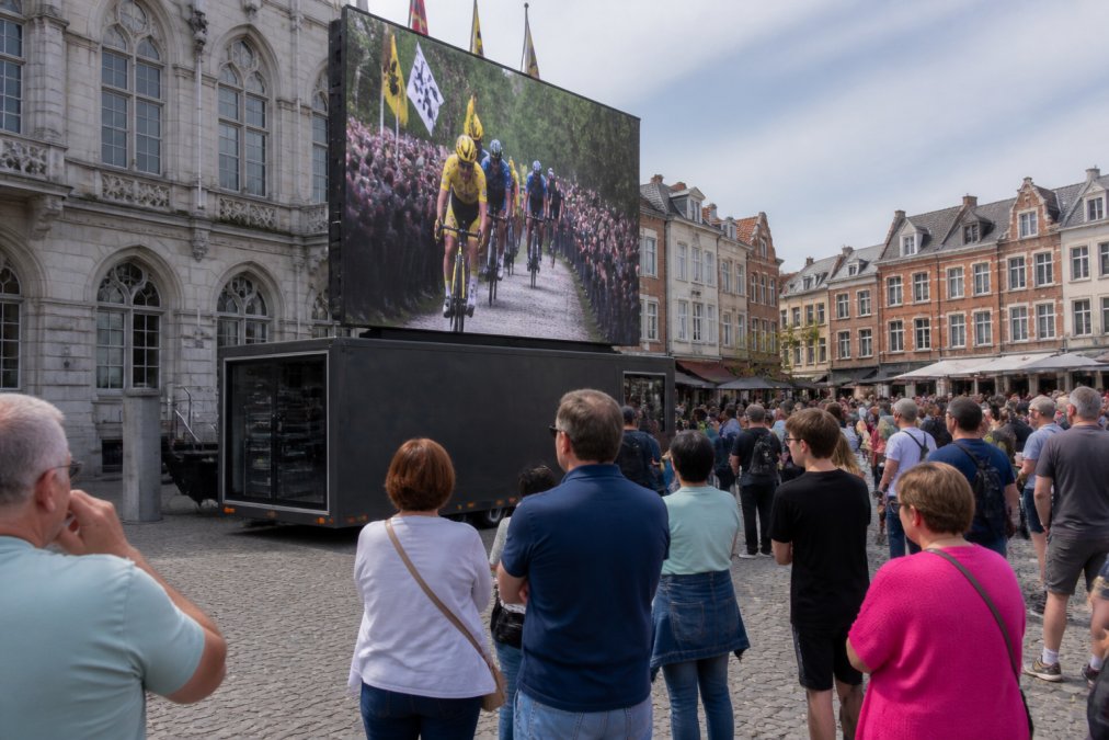 Een LED-scherm op een aanhangwagen tijdens een buitenmarketingevenement op een stadsplein in België, mensen kijken naar het heldere scherm, de aanhangwagen heeft een regieruimte, daglicht, realistische setting
