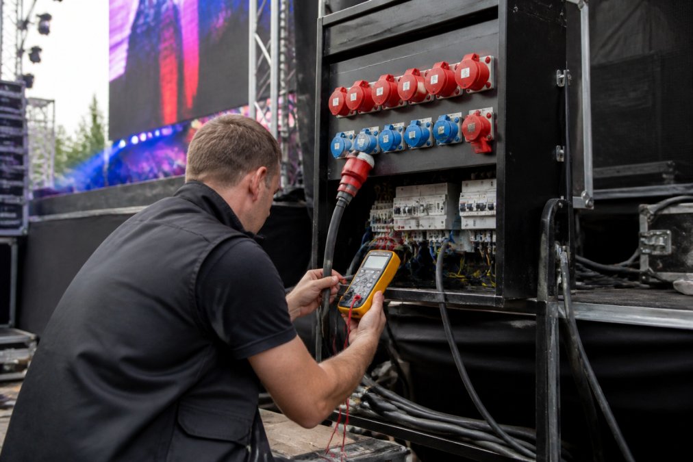 Professionele krachtstroom-verdeelkast op een evenement met LED-wand zichtbaar in de achtergrond, technicus controleert aansluitingen met digitale multimeter