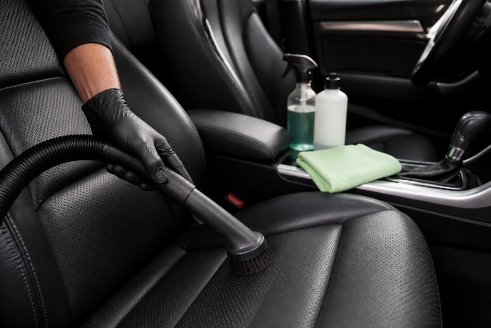 Close-up van iemand die een stofzuiger met zachte borstelkop gebruikt op leren autostoelen, met microvezeldoeken en reinigingsproducten zichtbaar op de passagiersstoel