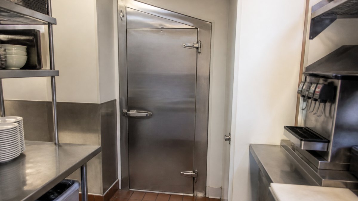 Restaurant kitchen with a walk-in cooler door installed in a tight corner space, showing stainless steel frame and prehung door fitted into an irregularly-shaped opening beside prep counters and shelving
