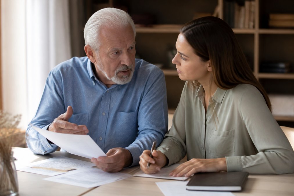 Adult child and parent sitting together at a desk with financial documents, having a serious conversation in a home office with warm lighting
