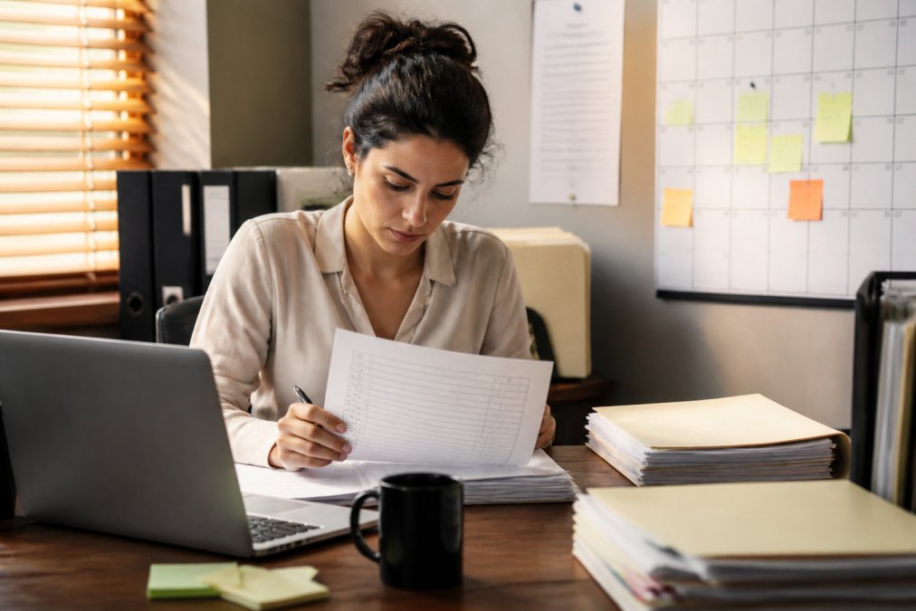 A paralegal at a tidy desk reviewing a printed case file index beside a laptop showing a document folder structure, sticky notes marking deadlines on a wall calendar, warm afternoon light through blinds