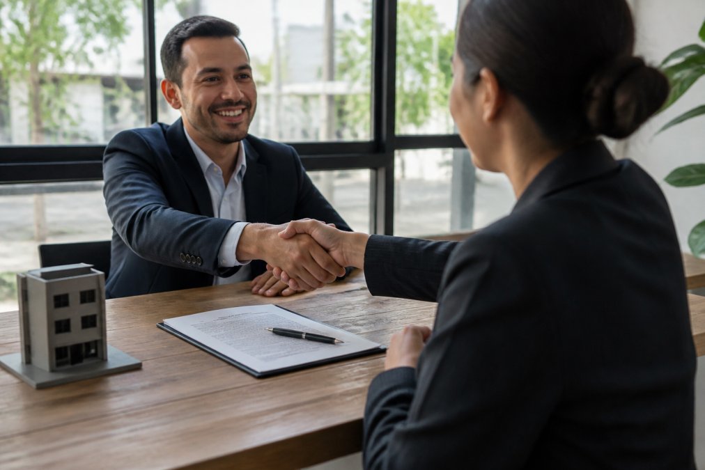 Two professionals in business attire shaking hands across a desk during lease negotiation meeting, with lease agreement and pen visible, natural window lighting in background