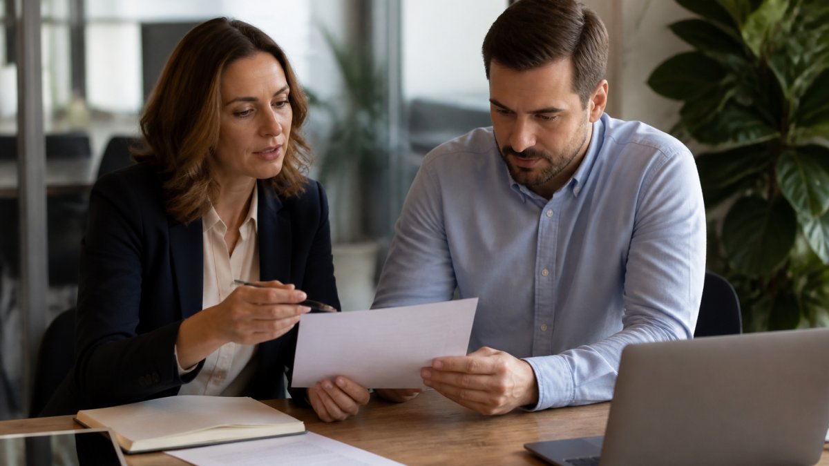 Marketing manager and legal advisor reviewing campaign documents and contract terms at a desk with laptop and tablet, natural office lighting