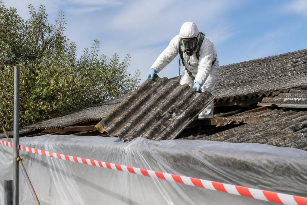 Worker in full protective suit and respirator carefully removing an old asbestos cement roof panel, with plastic sheeting covering the ground and warning tape around the work area