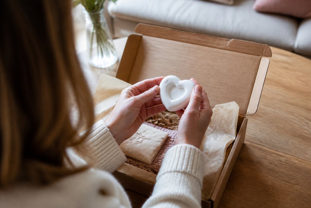 A person's hands holding a small, hand-finished keepsake gift that has just been taken out of a letterbox-sized parcel, with soft natural light and a warm home background