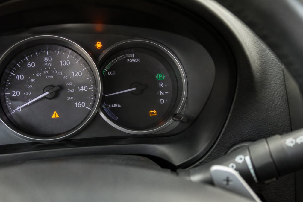 Close-up of a Lexus dashboard with hybrid system warning light illuminated in amber, showing the battery warning indicator clearly visible on the instrument cluster