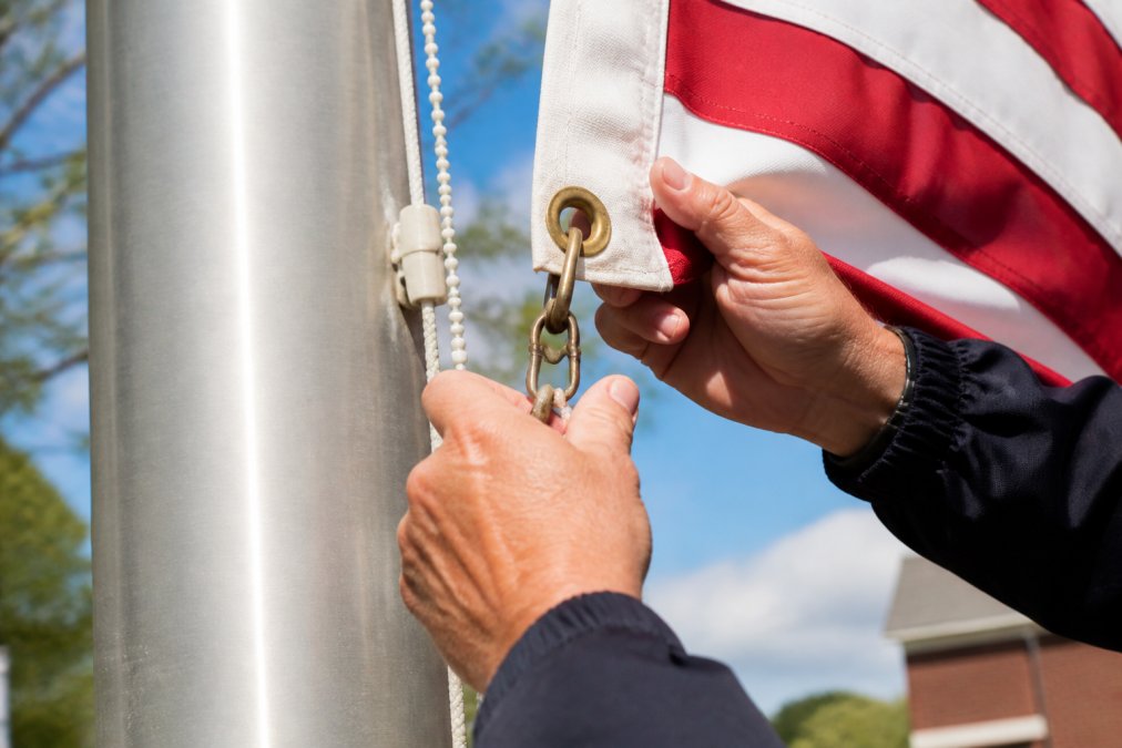 Close-up of flag hardware showing brass grommets, snap hooks, and halyard system being inspected on outdoor flag, with hands performing maintenance check in natural daylight