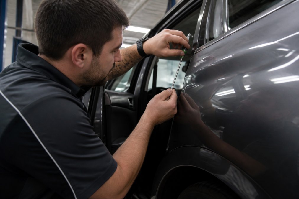 Professional technician performing paintless dent repair on hail-damaged vehicle using specialized metal-working tools in a modern repair facility with bright overhead lighting