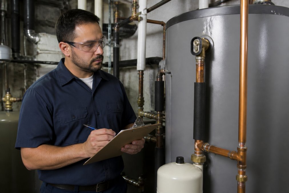 Licensed commercial plumber inspecting a large water heater in a mechanical room while wearing safety gear