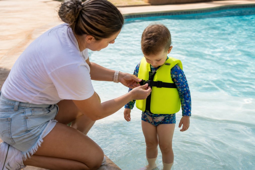 Parent checking fit of a toddler swim vest at poolside, demonstrating proper chest strap fastening and secure positioning with child standing in shallow water