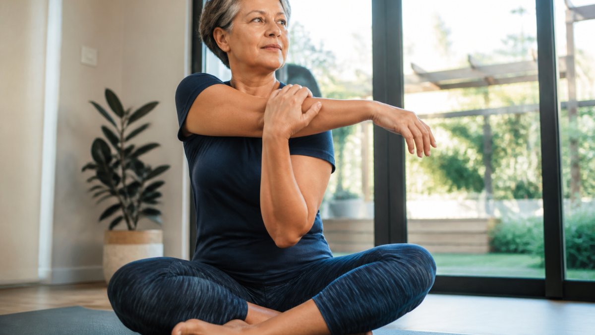 Middle-aged person performing gentle stretches on a yoga mat in a sunlit room as part of healthy lifestyle habits.