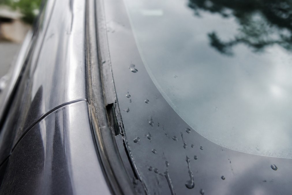 Close-up of a windshield showing visible water leaks seeping along the molding, air gaps around the edges, and reflective light highlighting improper seal areas and installation defects