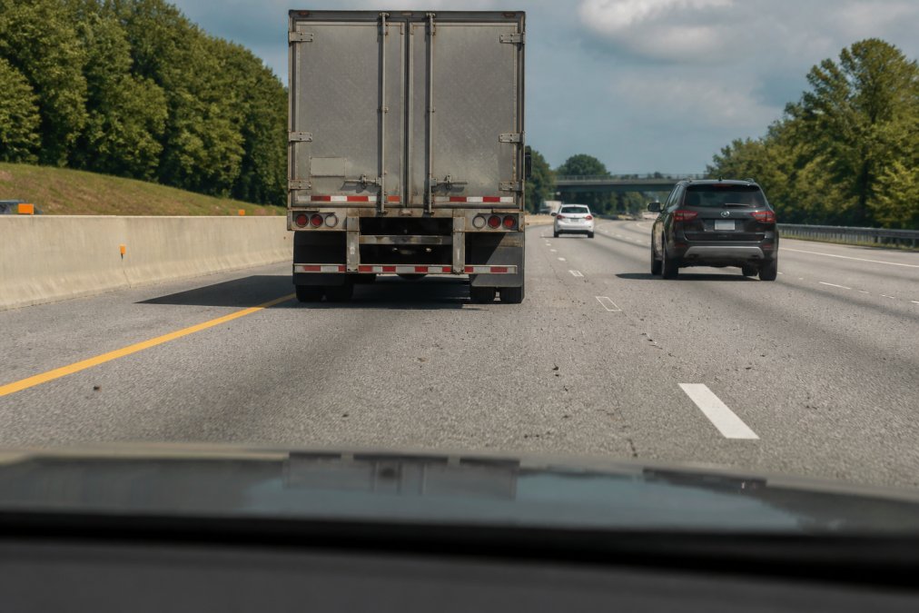 Driver maintaining safe distance behind a commercial truck on a multi-lane highway, with visible road debris and gravel particles on the asphalt, demonstrating defensive driving positioning