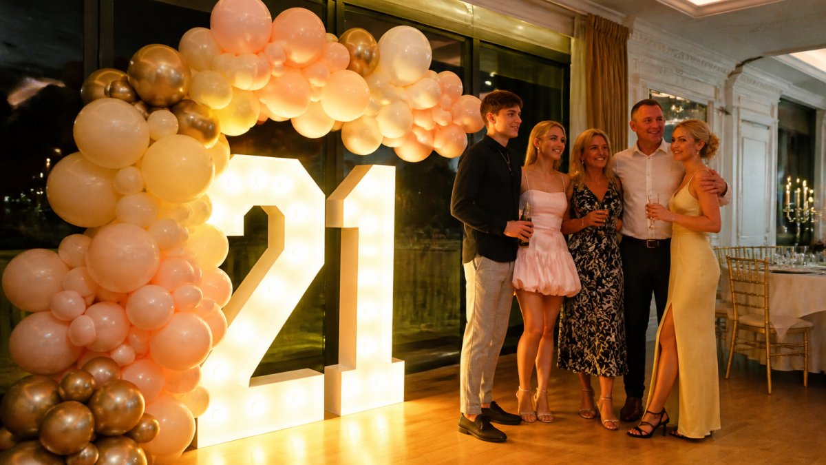 A 1.2m light-up number display glowing beside a pastel balloon arch in an elegant party venue, guests posing for photos in front of the illuminated digits under warm evening lighting