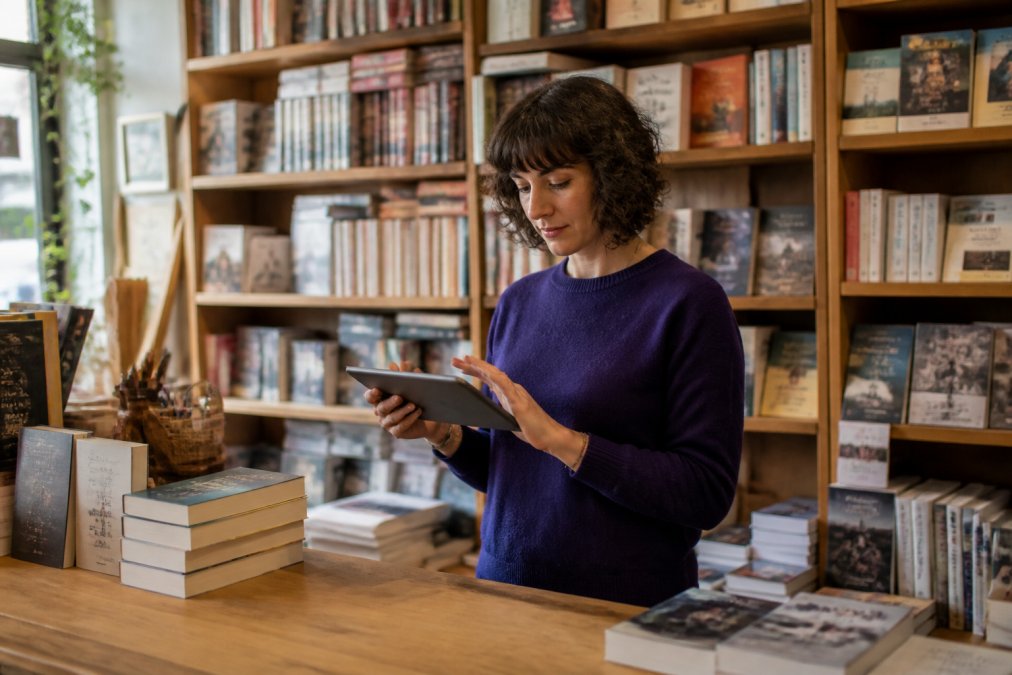 A bookseller at a wooden counter in an independent bookstore using a tablet to check inventory, shelves of books visible in the background, warm natural light